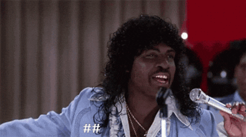 a man with long hair is singing into a microphone with a hashtag on his shirt