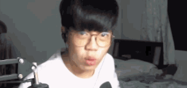 a young man wearing glasses and headphones is sitting in front of a microphone in a bedroom .