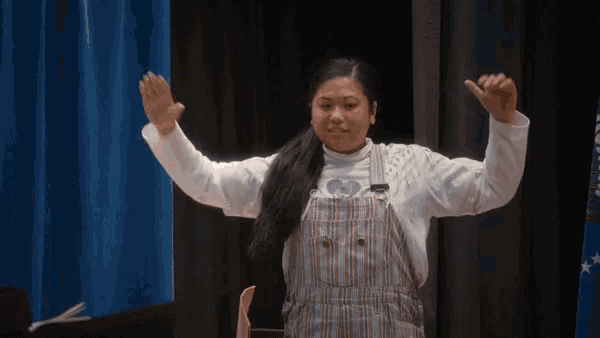 a girl in overalls is standing on a stage with her hands in the air