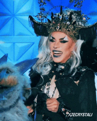 a drag queen with a crown on her head is smiling and holding a stuffed animal