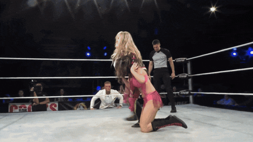 a woman in a pink bikini is kneeling in a wrestling ring while a referee watches