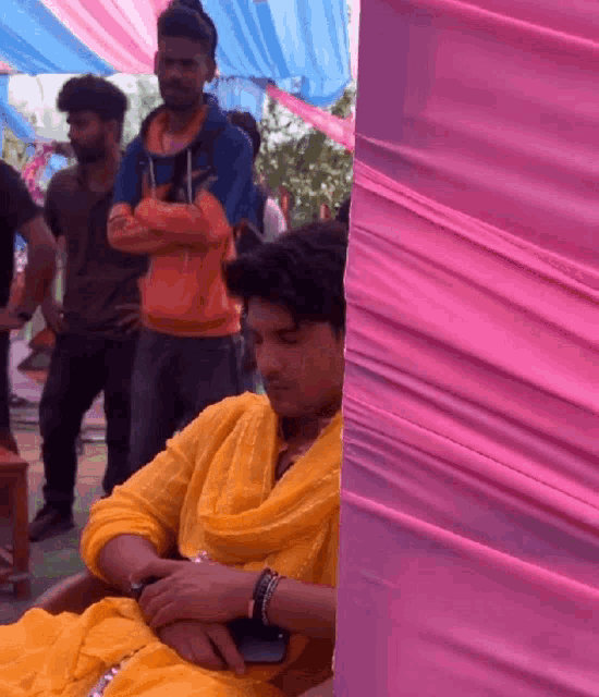 a man in a yellow dress is sitting in front of a pink curtain