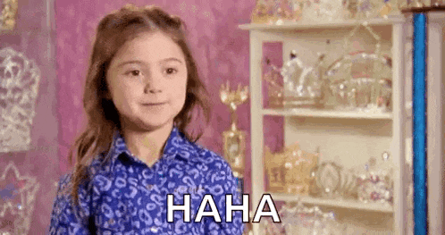 a little girl in a blue shirt is laughing in front of a shelf with trophies .