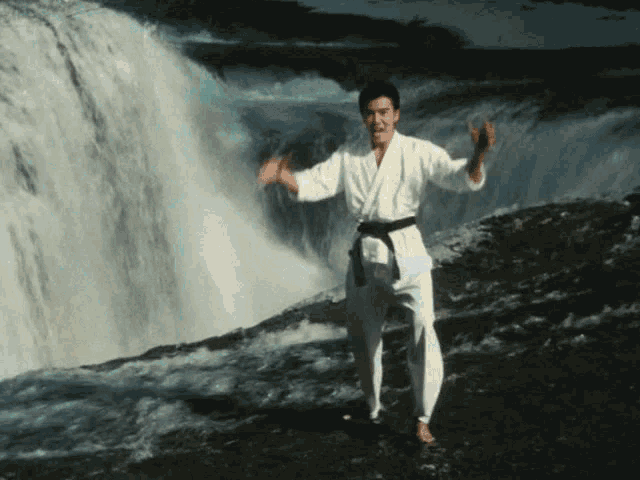 a man in a white karate uniform with a black belt stands in front of a waterfall