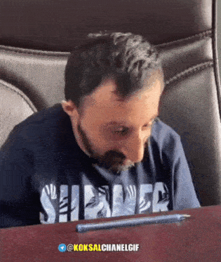a man sitting at a table with a pen in front of him and a shirt that says shiner