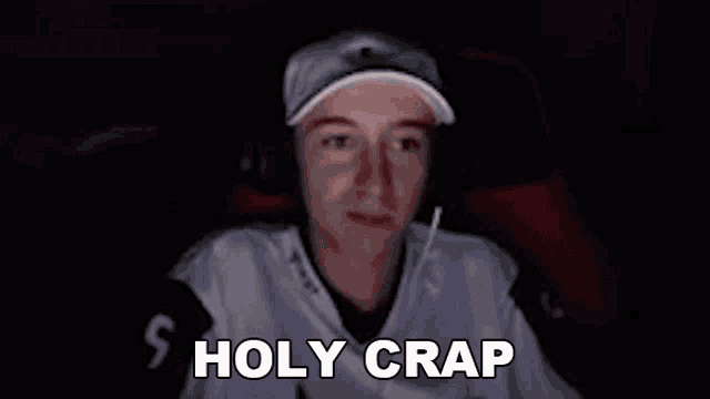 a young man wearing headphones and a hat is sitting in front of a computer screen and says `` holy crap '' .
