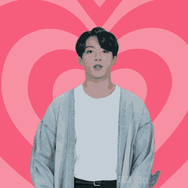 a man wearing a grey cardigan is standing in front of a pink heart shaped background