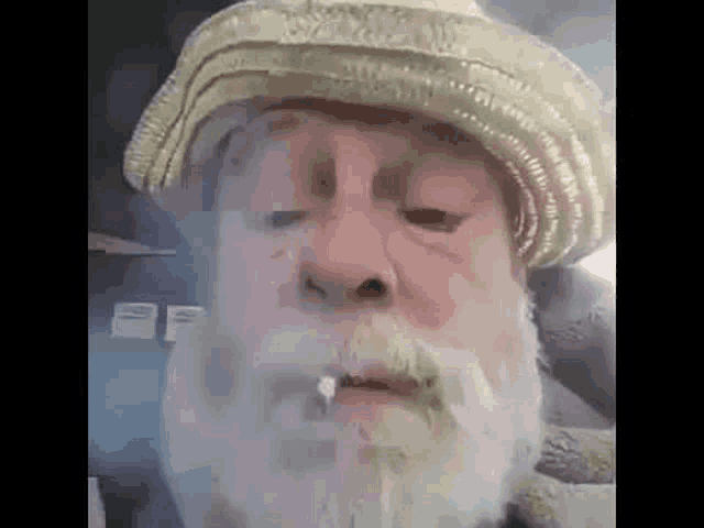 an elderly man with a beard and hat is smoking a cigarette .