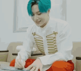a young man with blue hair is sitting on a couch writing on a piece of paper with a pen .