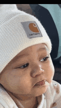 a baby wearing a white carhartt beanie is looking at the camera with his mouth open .