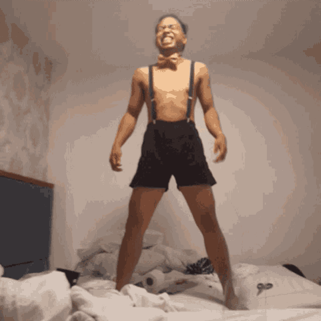 a shirtless man with suspenders and a bow tie is standing on a bed