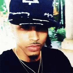 a man wearing a hat and a necklace is looking at the camera .