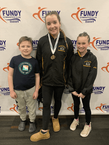 a girl with a medal is standing next to two other children in front of a fundy gymnastics club banner