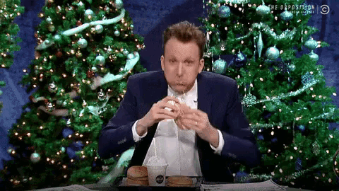 a man in a suit is eating a hamburger with a straw in front of a christmas tree