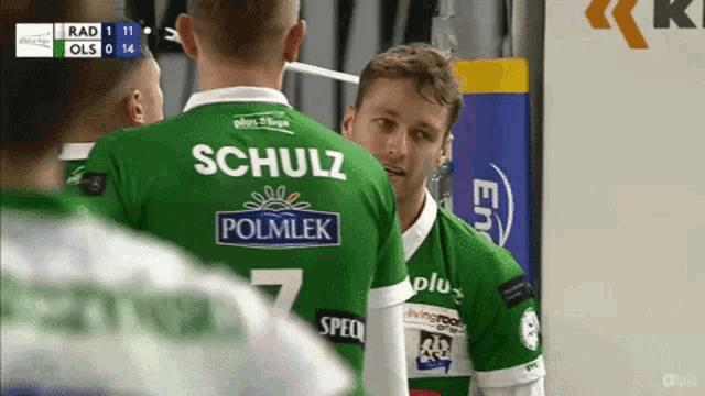 a man wearing a green schulz polmlek jersey talks to another man