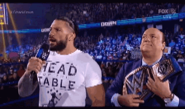two men are standing next to each other in front of a microphone . one of the men is wearing a lead table shirt .