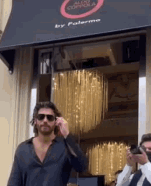a man wearing sunglasses is talking on a cell phone in front of a building that says aldo confola by palermo