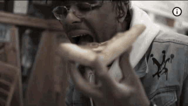 a man wearing glasses and a denim jacket is eating a sandwich