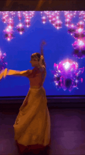 a woman in a long yellow dress is dancing in front of a large screen