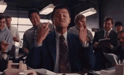 a man in a suit and tie is sitting at a desk in front of a group of people and giving the middle finger .