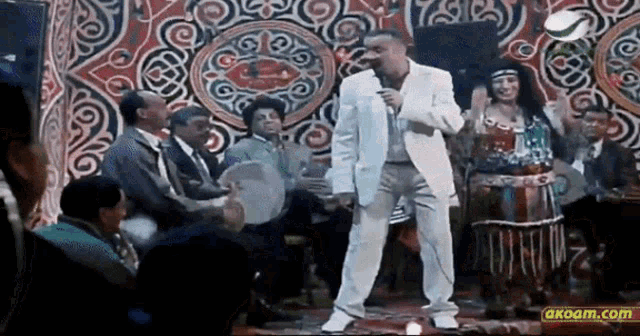 a man in a white suit is dancing in front of a crowd with a akoam.com logo