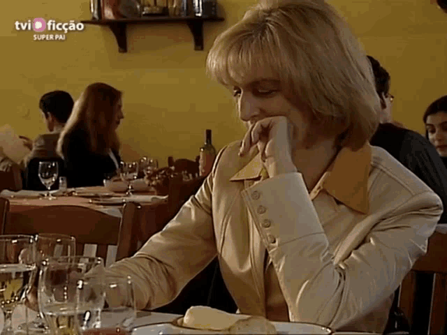 a woman sits at a table in a restaurant with tvi ficcao super pai written on the bottom right