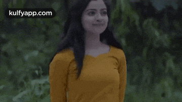 a woman in a yellow shirt is standing in front of a forest and smiling .