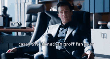 a man in a suit sits in a chair with the words casey the johnathan groff fan over written below him