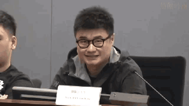 a man wearing glasses and a black jacket is sitting at a table with a microphone .