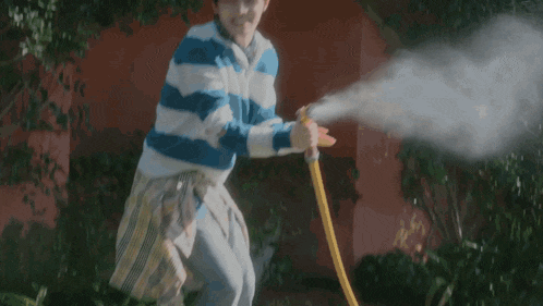 a man in a blue and white striped shirt is watering plants with a yellow hose