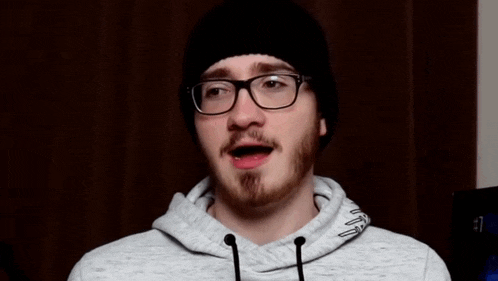a man with glasses and a beard is singing into a microphone while wearing a beanie and a hoodie .