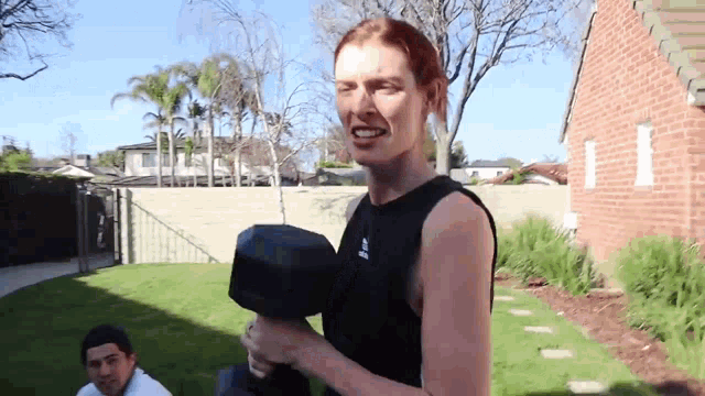 a woman is holding a dumbbell in a backyard while a man sits on the grass .