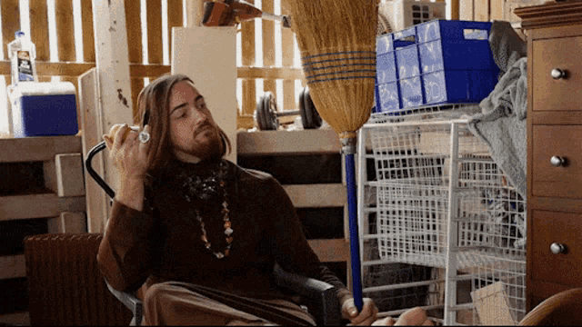 a man with long hair sits in a chair with a broom in front of him and a blue cooler