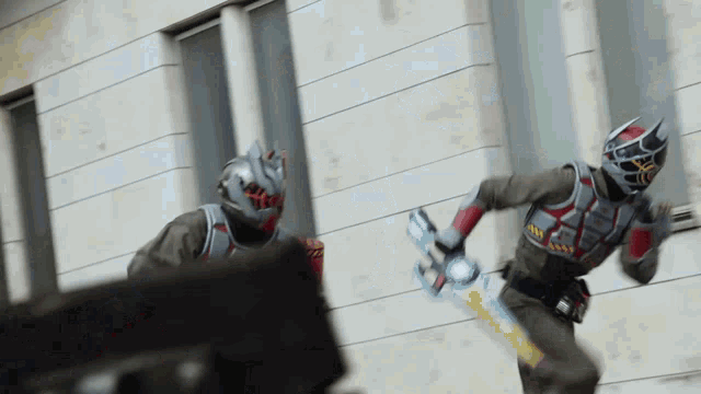 two men in superhero costumes are fighting in front of a building