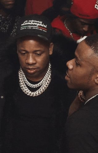 a man wearing a lamborghini hat talks to another man in a crowd