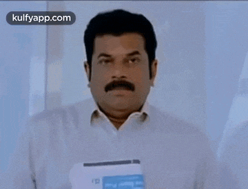 a man with a mustache is standing in front of a white wall holding a piece of paper .