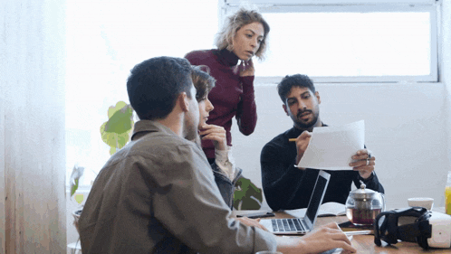 a group of people are sitting around a table with laptops and papers