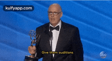 a man in a tuxedo is holding a trophy and saying give them auditions .