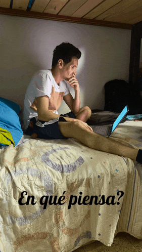a man sits on a bed looking at a laptop with the words en que piensa written below him