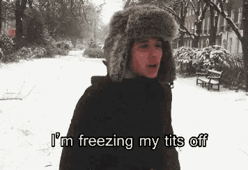 a man in a fur hat is standing in the snow and says i 'm freezing my tits off .