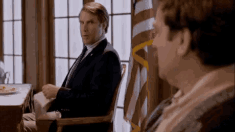 two men are sitting in chairs in front of a flag .