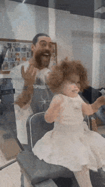 a little girl in a white dress is sitting next to a man with a beard in a barber shop .