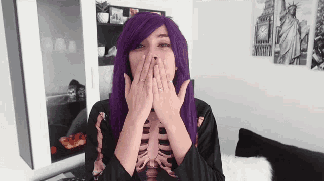 a woman with purple hair and a skeleton shirt is covering her mouth with her hands