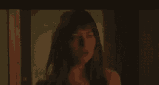 a woman with long hair is standing in a dark room looking out a window .