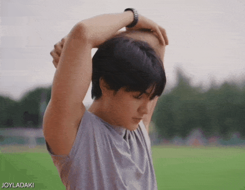 a young man is stretching his arms in front of a green field with joyladaki written below him