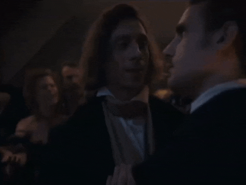 a man in a tuxedo and bow tie talks to another man in a dark room