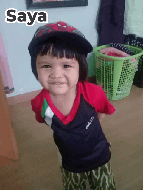 a little girl wearing a spiderman hat stands in front of a laundry basket