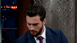 a man with a beard wearing a suit and tie is sitting in front of a window .