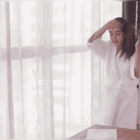 a woman in a bathrobe is standing in front of a window and brushing her hair .