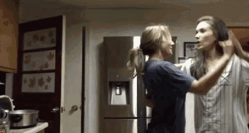 two women are standing in a kitchen and one of them is holding the other 's hair .
